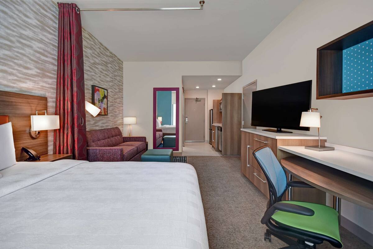 A hotel room with a large bed, a desk with a TV, a chair, a sofa, and a bathroom visible through an open door.