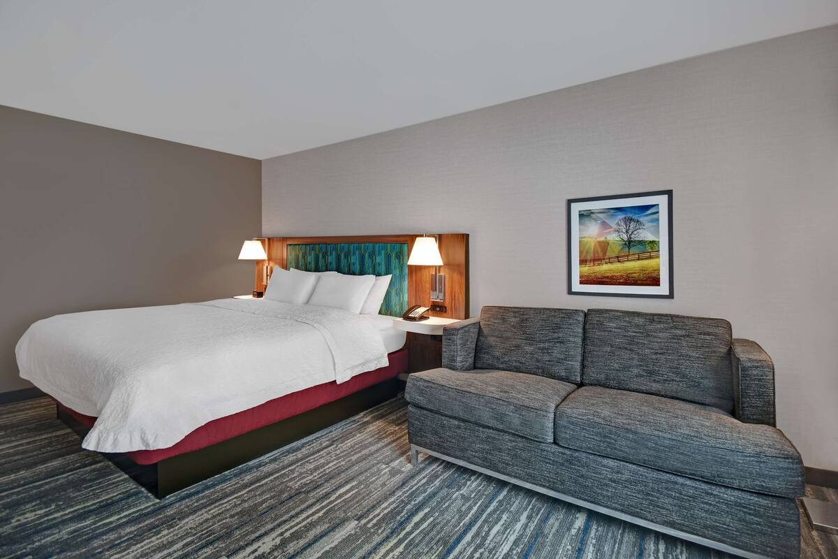A hotel room with a bed, a grey sofa, a nightstand with lamps, and a framed picture on the wall.