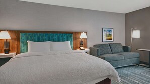 A hotel room with a large bed, two bedside tables, a grey sofa, a small table, and a painting on the wall.