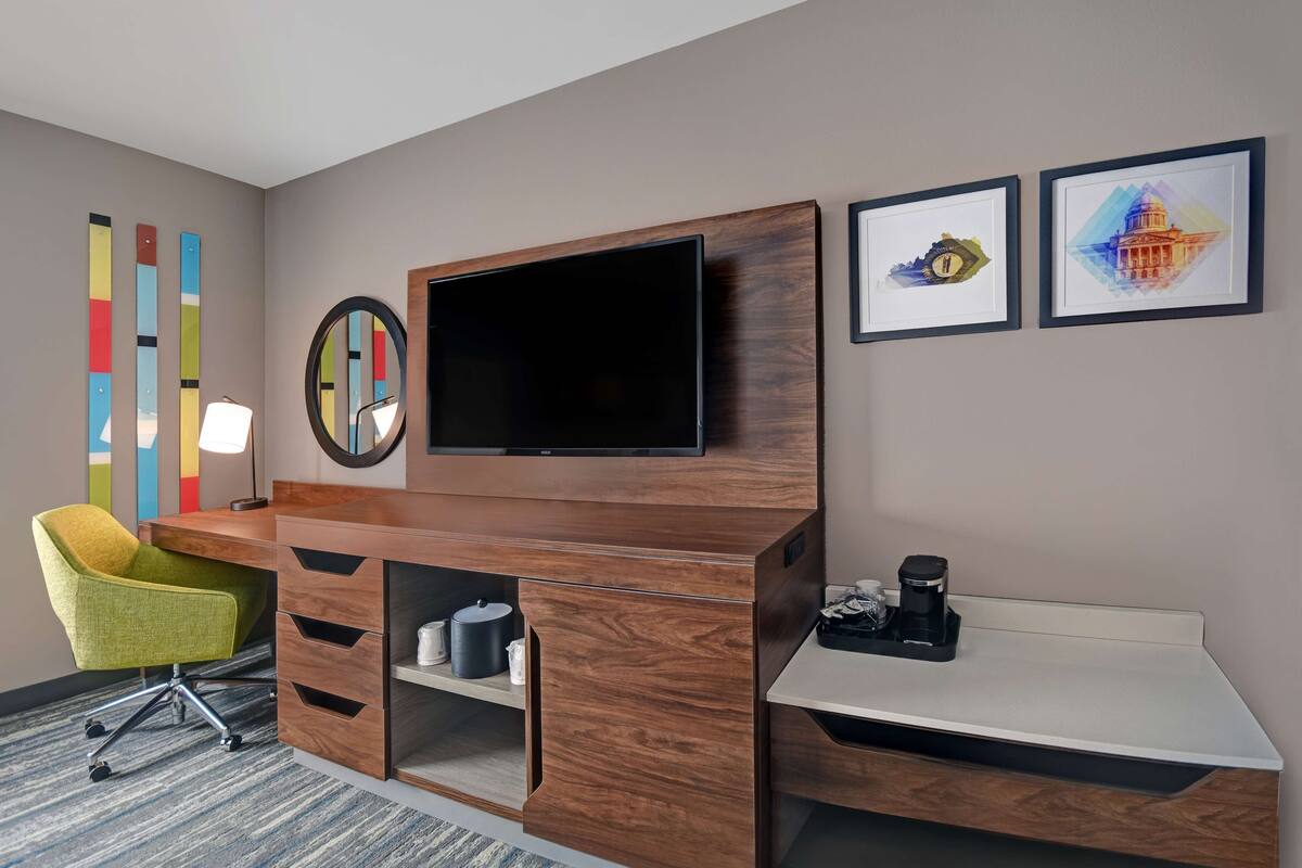 A hotel room with a wooden desk, a flat-screen TV, a green chair, and two framed pictures on the wall.