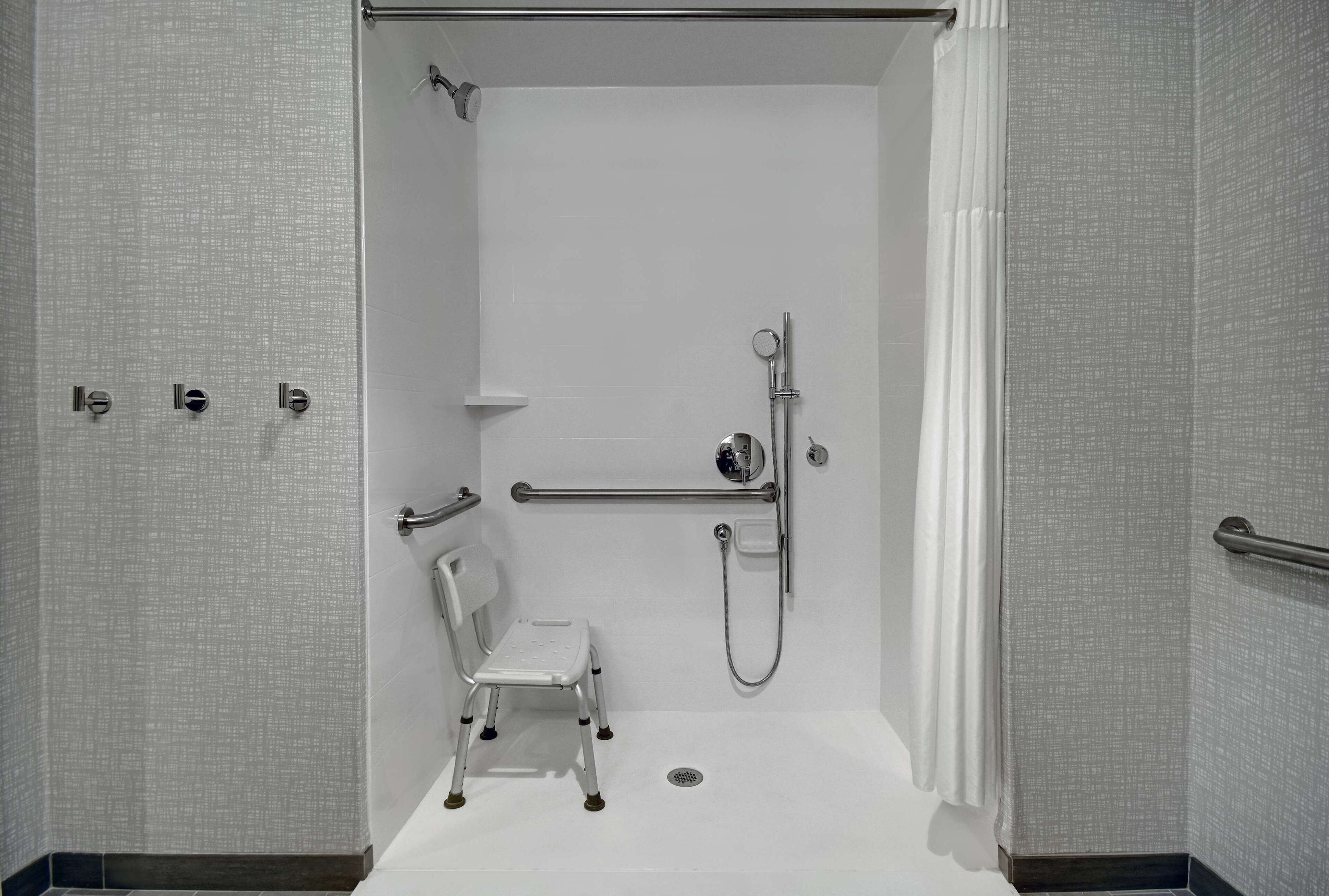 A small, white, walk-in shower with a handheld showerhead, grab bars, and a white chair.