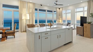 Une cuisine moderne dotée d’un grand îlot central, un coin repas offrant une vue sur la mer et un espace de vie avec un canapé et un téléviseur.