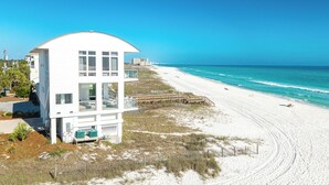 Une maison blanche située en bord de mer, dotée d’un toit incurvé, d’une terrasse en bois et d’une plage de sable en arrière-plan.