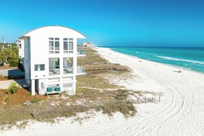 Una casa de playa blanca con techo curvo, una terraza de madera y una playa arenosa al fondo.