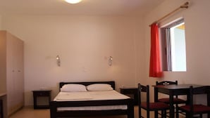 Ein Schlafzimmer mit einem Bett, Nachttischen, einem Schreibtisch mit Stühlen und einem Fenster mit roten Vorhängen.