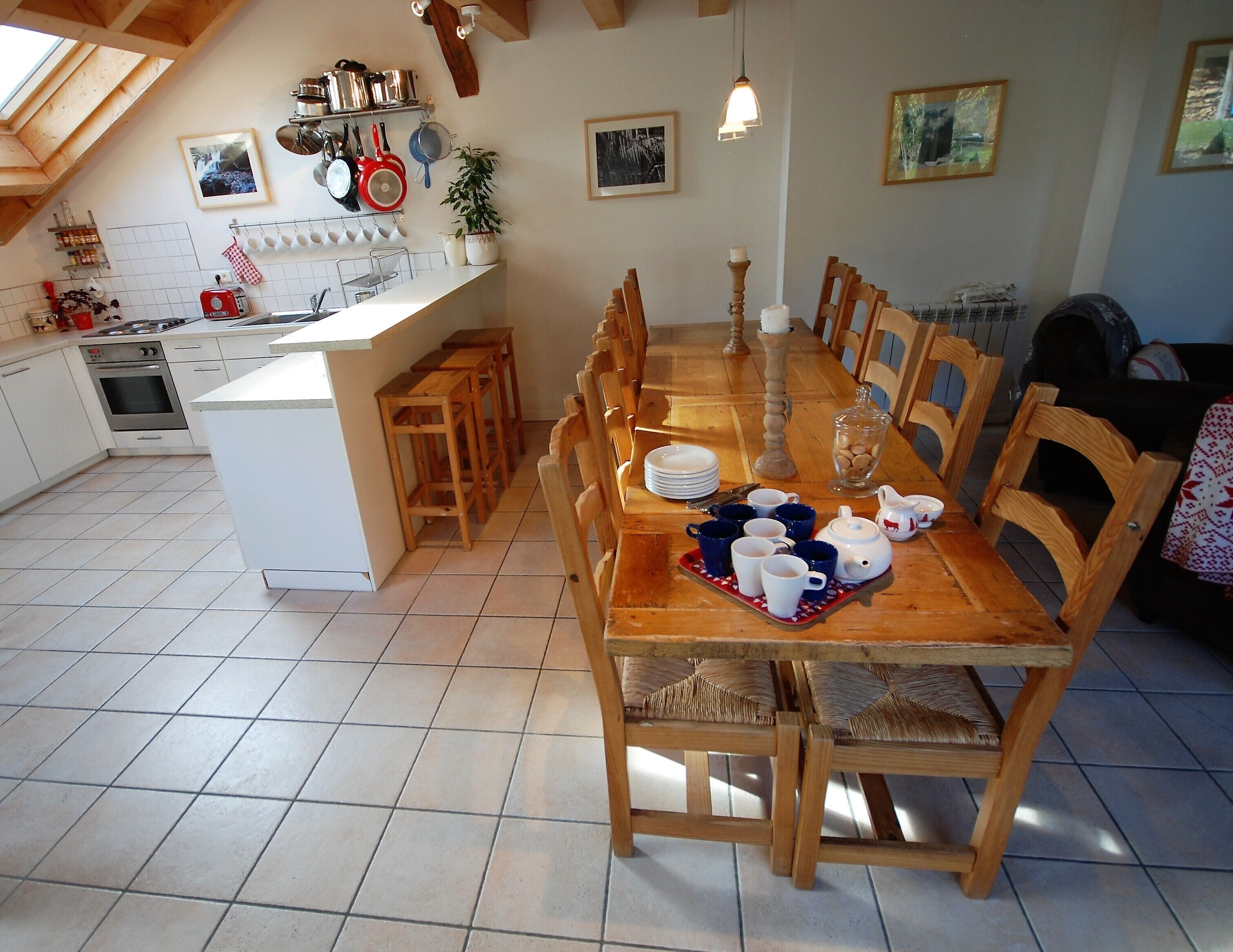 A dining area with a wooden table set for six, surrounded by chairs. A kitchen with white cabinets and a tiled backsplash is visible in the background.