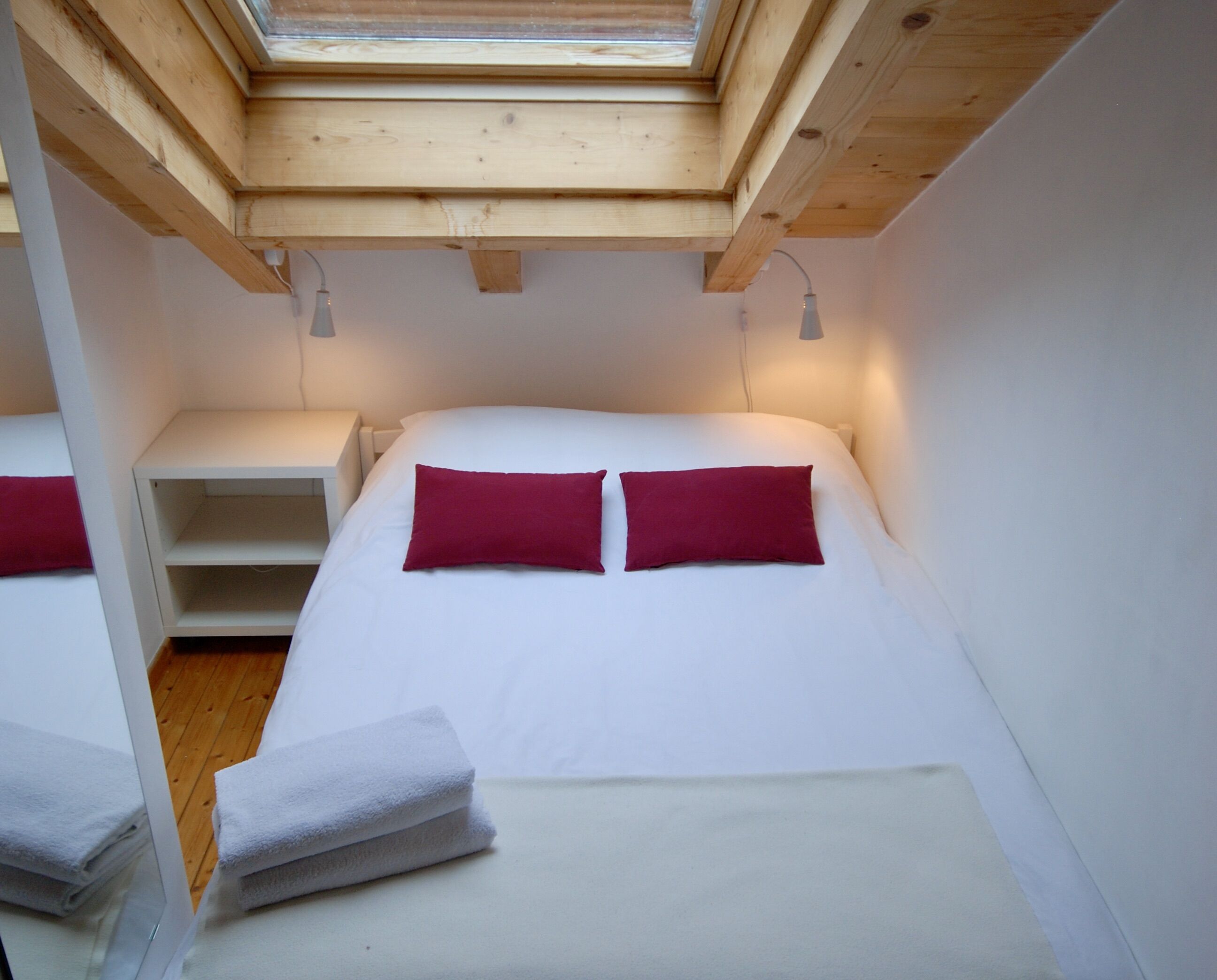 A bedroom with a skylight, a bed with white bedding and two red pillows, a bedside table, and a stack of white towels.