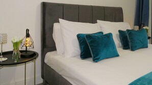 Ein ordentlich bezogenes Bett mit weißen und türkisblauen Kissen, ein Nachttisch mit Blumen in einer Vase und einer Lampe, sowie ein gerahmtes Bild über dem Bett.