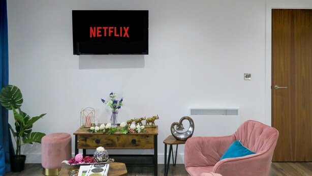 Ein Wohnzimmer mit einem Flachbildfernseher, der "NETFLIX" anzeigt, einem Holztisch mit einer Zeitschrift, einem rosa Sessel, einer Topfpflanze und einer Holztür.