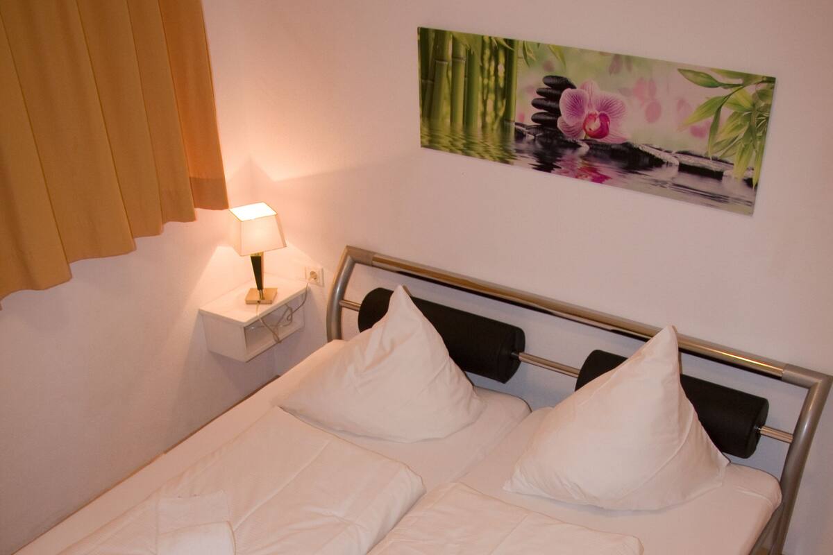Un lit jumeau bien fait, avec du linge de lit et des oreillers blancs, une table de chevet avec une lampe, et un tableau représentant des fleurs et du bambou accroché au mur.