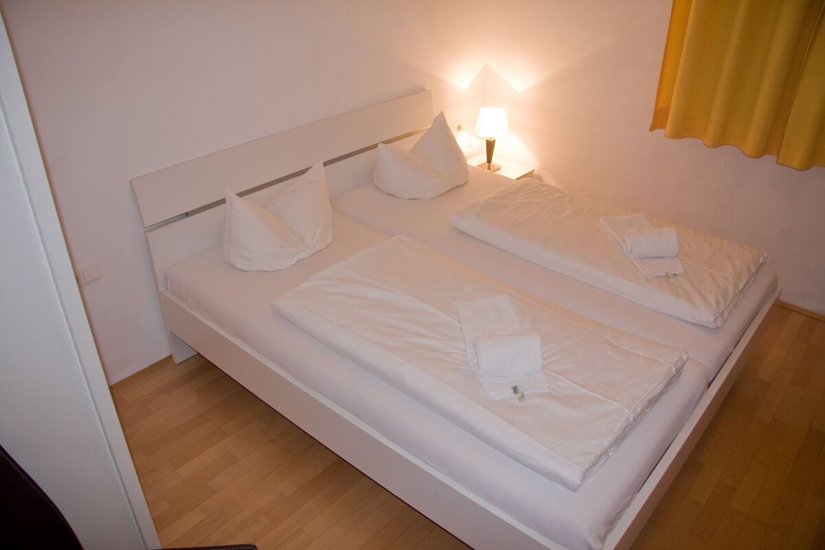 Un lit double avec du linge de lit blanc et des oreillers, une lampe de chevet et un plancher en bois.