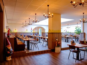 Un'ampia sala ristorante con soffitti alti, diversi lampadari, tavoli e sedie in legno e statuette decorative a forma di gnomi.