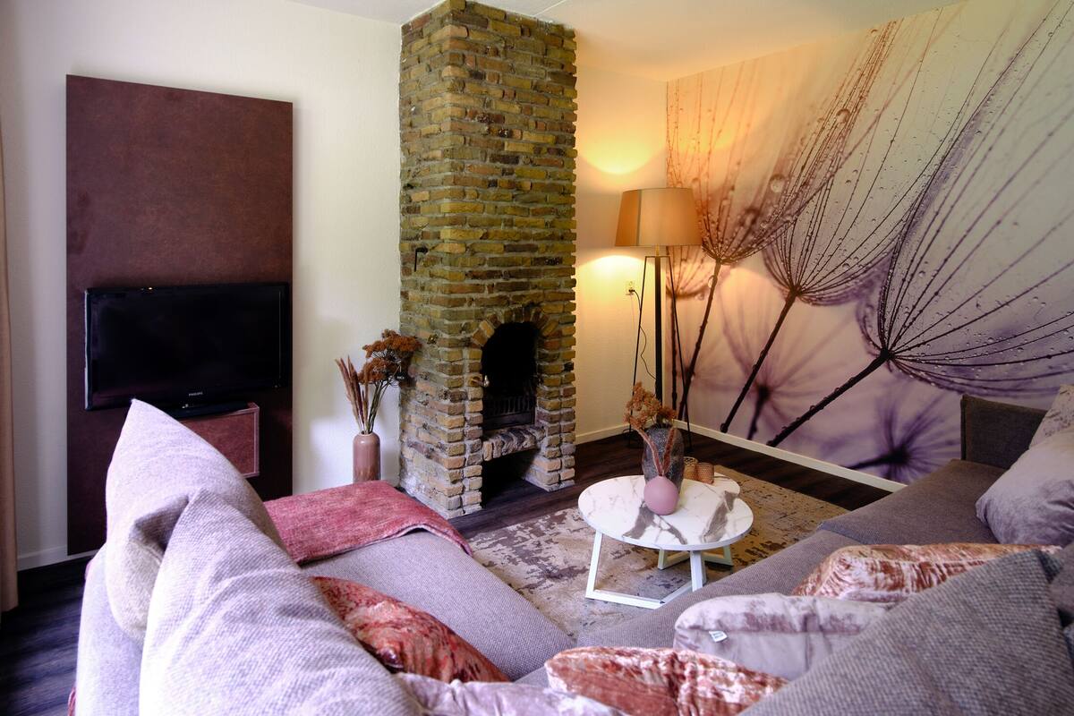 A living room with a brick fireplace, a flat-screen TV, a sofa with cushions, and a decorative plant.