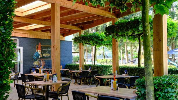 Un patio al aire libre con mesas y sillas de madera, un mural en la pared y abundante vegetación alrededor.