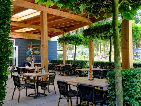 Een terras buiten met houten tafels en stoelen, een muurschildering en begroeiing rondom.