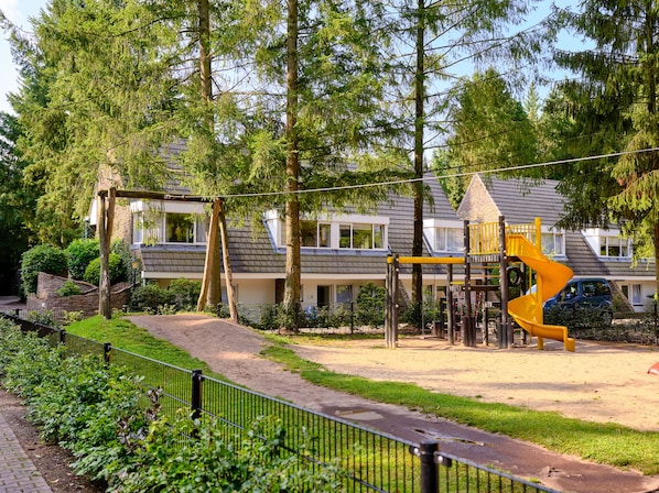 A playground with a yellow slide and swings, surrounded by trees and a building with a sloped roof. - Vakantiepark De Bosrand (Vaassen)