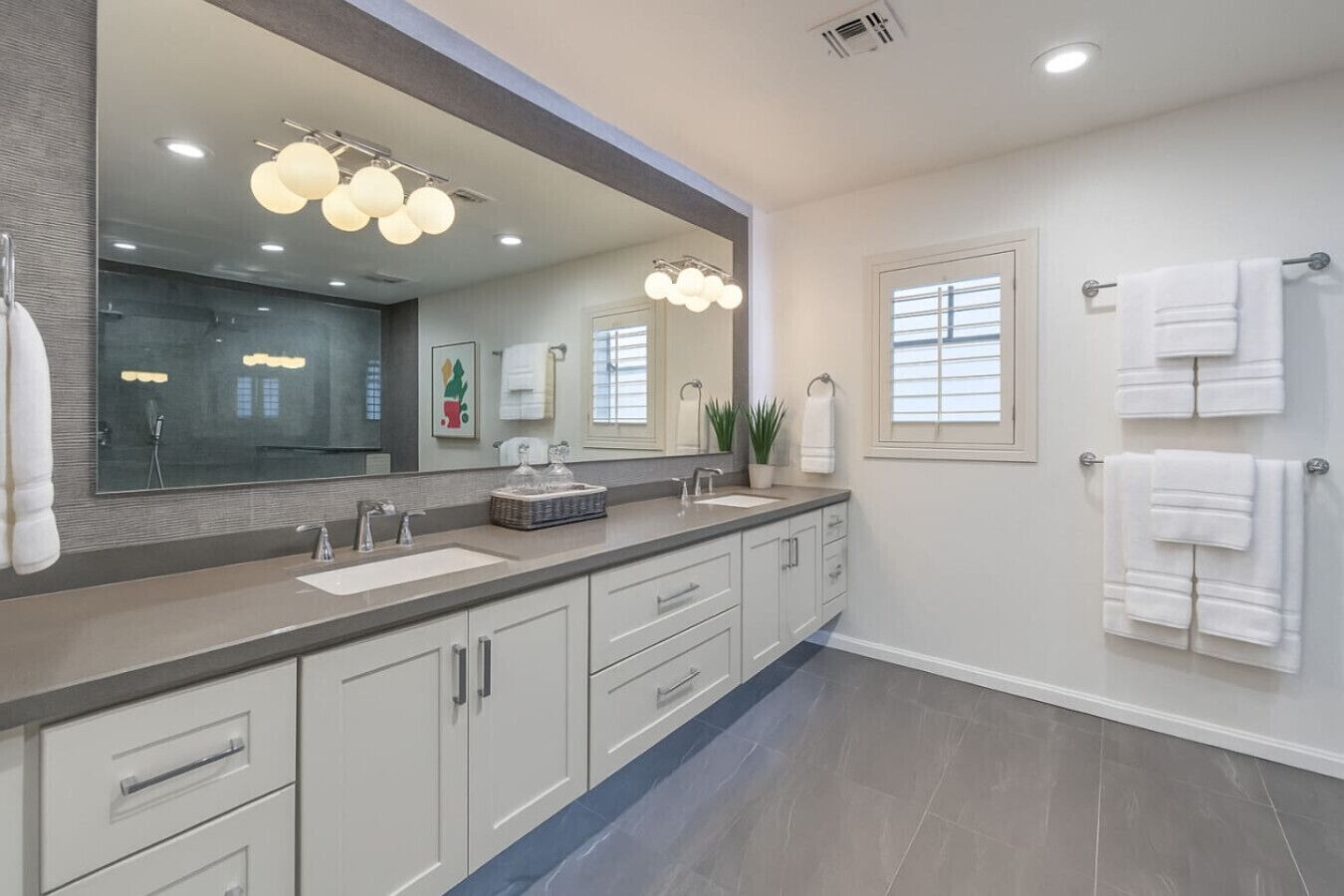 A modern bathroom with a large mirror, two sinks, and a vanity with drawers. There are towels hanging on a rack and a potted plant on the vanity.