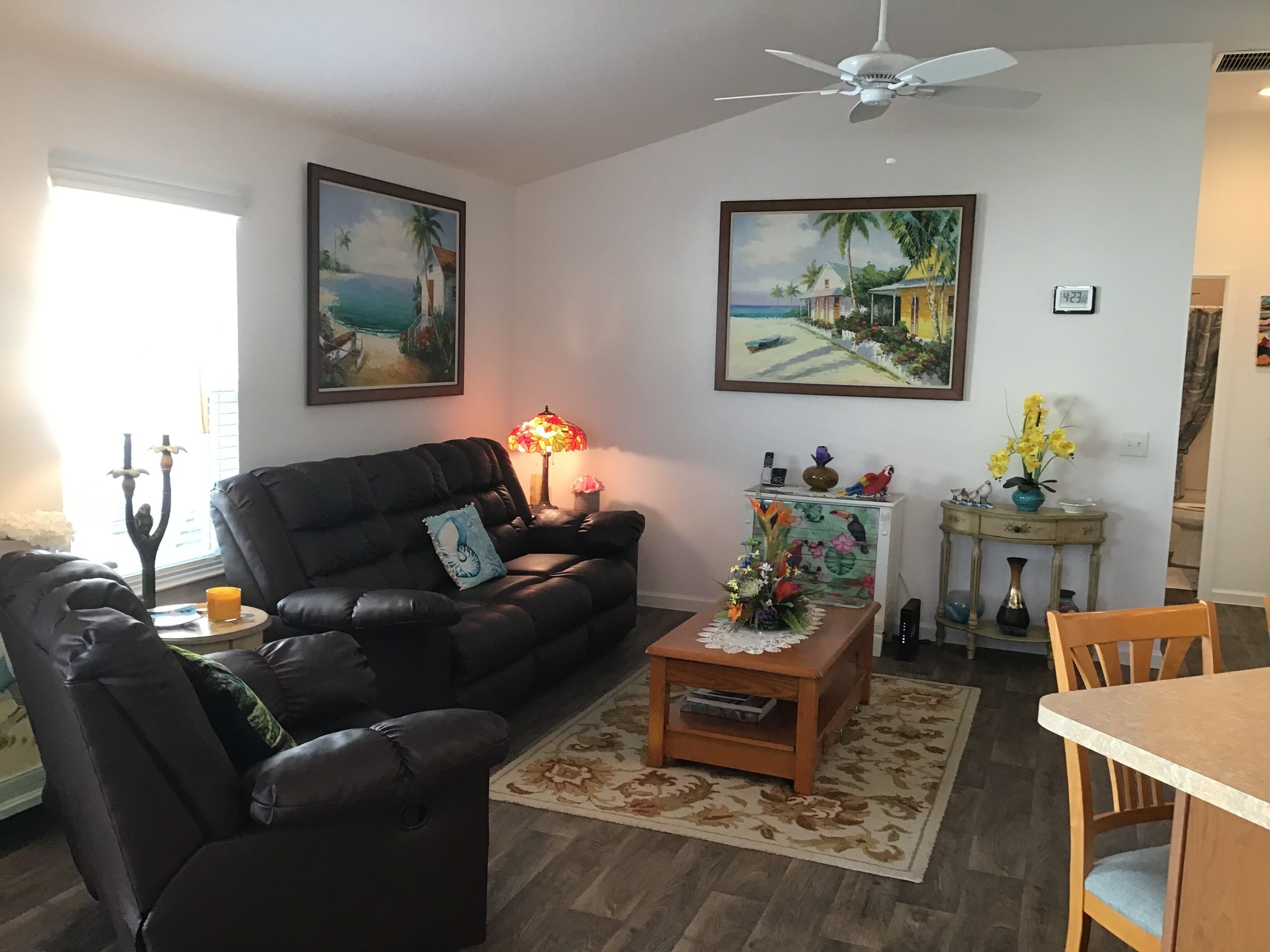A living room with a dark leather sofa, a wooden coffee table, and a dining table with chairs. There are paintings on the walls, a lamp on the coffee table, and a ceiling fan.