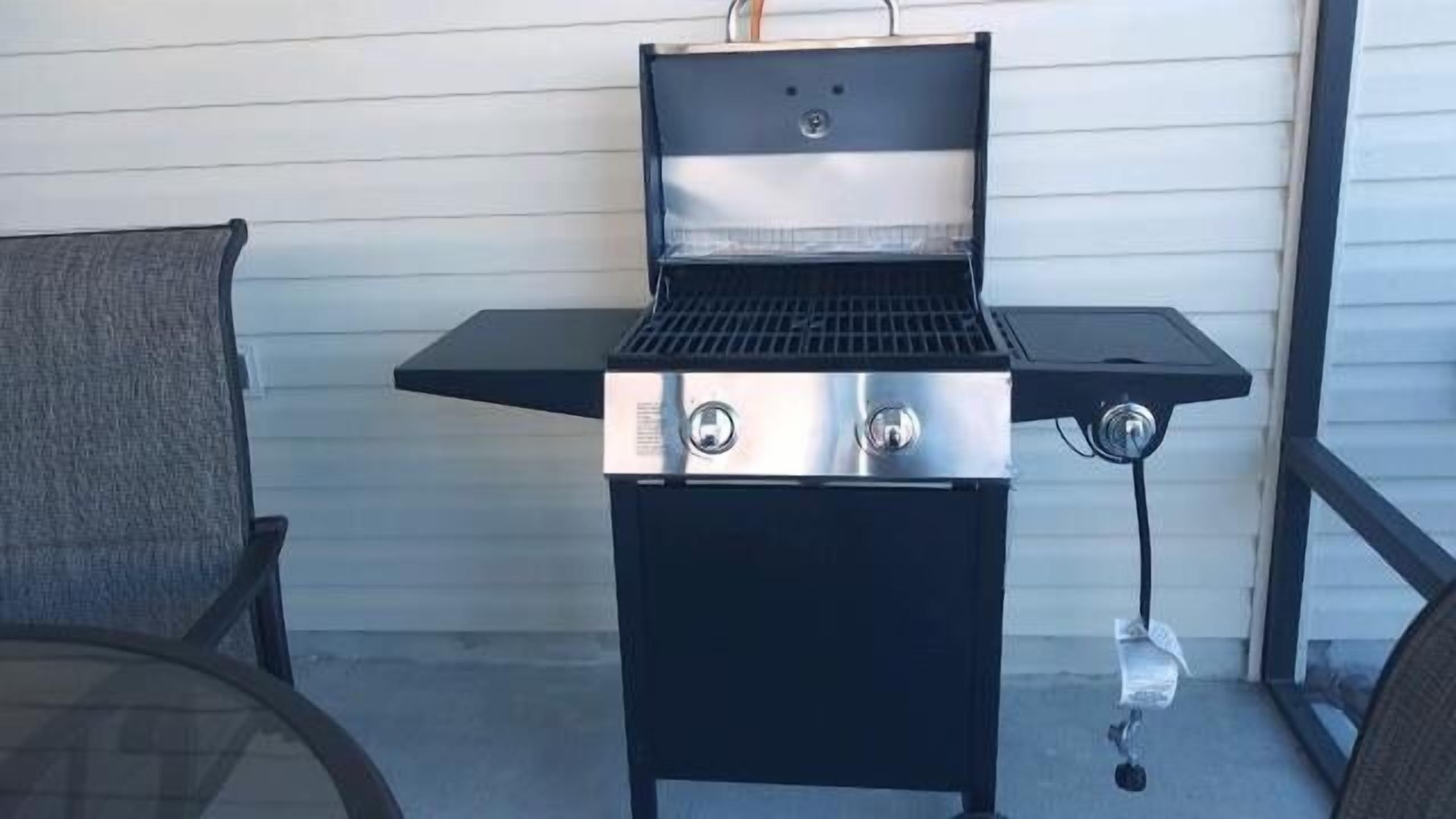 A portable grill with a control panel and a gas hose, placed on a patio with a chair and a white wall in the background.