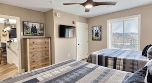 A bedroom with two beds, a wooden dresser, a TV, and a city view through the window. - Downtown Gatlinburg Luxury Penthouse, Mountain Views & 2 Balconies (Gatlinburg)