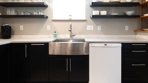 Een moderne keuken met donkere kastjes, een RVS spoelbak en een vaatwasser. Boven de spoelbak staan planken met glaswerk en servies.