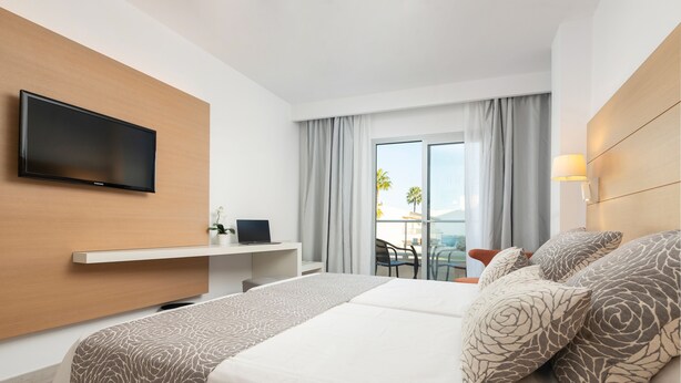 Quarto de hotel com cama, televisão na parede, secretária com computador e vista para o exterior através de uma grande janela.