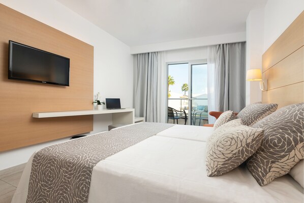 Quarto de hotel com cama, televisão na parede, escrivaninha com computador e varanda com vista.