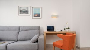 Uma sala de estar moderna com um sofá cinzento, uma escrivaninha de madeira com um laptop e uma cadeira laranja. Há duas imagens emolduradas na parede.
