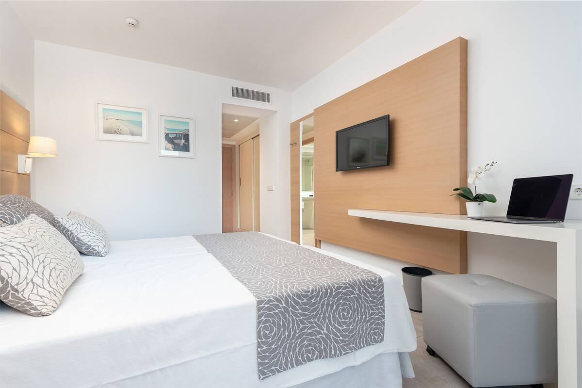 Une chambre d’hôtel moderne avec un lit bien fait, un téléviseur fixé au mur, un petit bureau avec un ordinateur portable et une plante en pot.