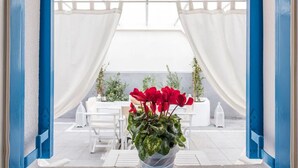 A view through a blue-framed window into a patio area with white curtains, a table, chairs, and potted plants.
