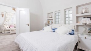 A bedroom with a large bed, white walls, and a white door. There is a white dresser with a mirror and a small table with a lamp.