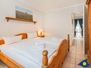 Un lit bien fait, avec du linge de lit et des serviettes blanches, une tête de lit en bois, une lampe de chevet et un tableau représentant un champ accroché au mur.