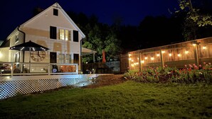 Ein zweistöckiges Haus mit einer großen Uhr an der Hauswand, einer überdachten Terrasse mit Sitzgelegenheiten und einem Garten mit roten Blumen.