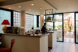 Un moderno vestíbulo de hotel con una recepción, un amplio ventanal con vista al área de estar exterior y un bar con botellas.