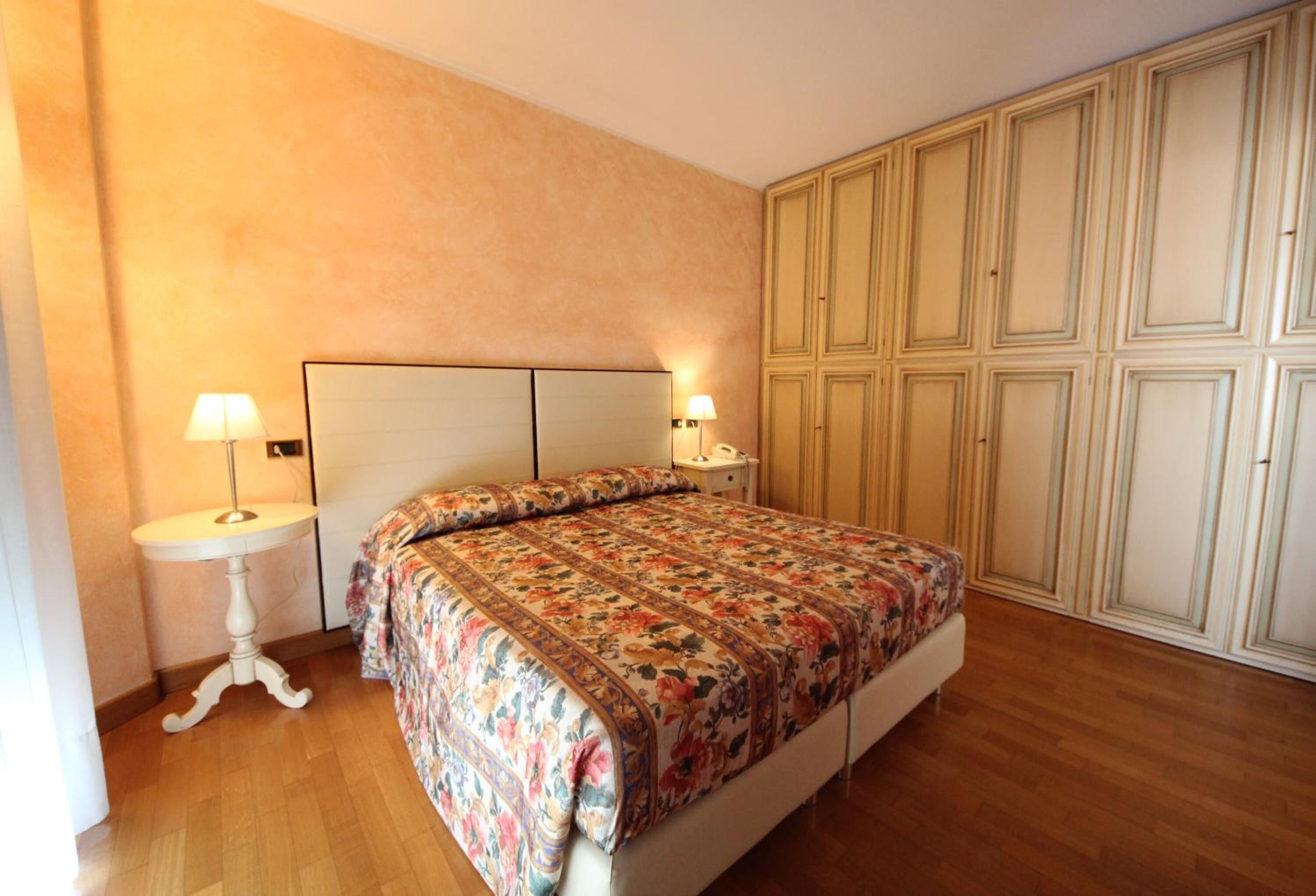 A bedroom with a bed, two bedside tables with lamps, wooden flooring, and a wardrobe with multiple doors.