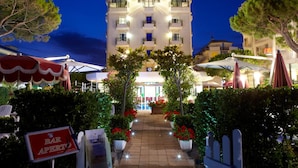 Entrada de hotel con un arco central, rodeada de plantas en macetas y mobiliario exterior.