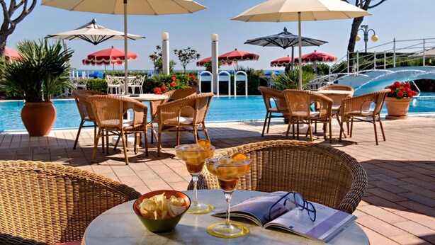 Zona junto a la piscina con sillas de mimbre, sombrillas y una mesa con bebidas y aperitivos.
