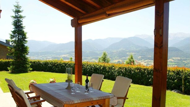 Une terrasse extérieure avec une table et des chaises en bois, une bouteille de vin et un verre. La terrasse offre une vue sur un jardin luxuriant et des montagnes lointaines.