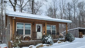 A wooden cabin with a snow-covered roof, large windows, and a front porch with a bench and a table. - Cozy cottage on our private lake and 75 wooded acres. (Dunlap)