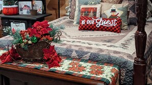 A cozy, well-decorated bedroom with a wooden headboard, a large bed with a quilted bedspread, a bedside table with a lamp, a small Christmas tree, and a framed picture on the wall. - Cozy cottage on our private lake and 75 wooded acres. (Dunlap)