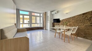 A modern living room with a large window, a stone accent wall, and a dining area with white furniture. - Agi Pi Sunyer 4 2D Apartamento for rent in Roses Girona. (Roses)