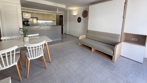 A modern living room with a dining area, a sofa, a kitchen in the background, and a large window. - Agi Pi Sunyer 4 2D Apartamento for rent in Roses Girona. (Roses)
