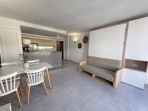 En moderne stue med spiseplads, sofa og et køkken i baggrunden, samt et stort vindue.