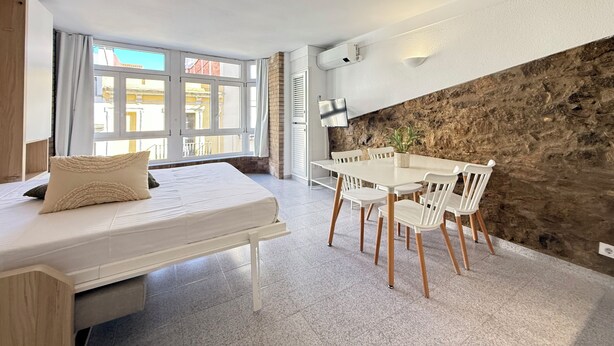 A bedroom with a bed, a dining table with chairs, a TV, and a stone wall. - Agi Pi Sunyer 4 2D Apartamento for rent in Roses Girona. (Roses)
