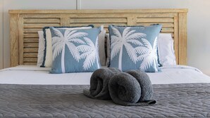 A bed with a wooden headboard, two pillows with a palm leaf design, and a rolled-up towel on a gray quilted bedspread. - Ocean View 49A Wi-Fi Pool King bed (Airlie Beach)
