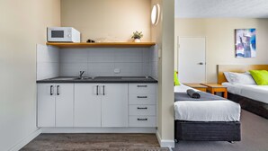 Ein kompaktes Hotelzimmer mit Küchenzeile, einem Bett mit grauem Kopfteil und einem kleinen Nachttisch.