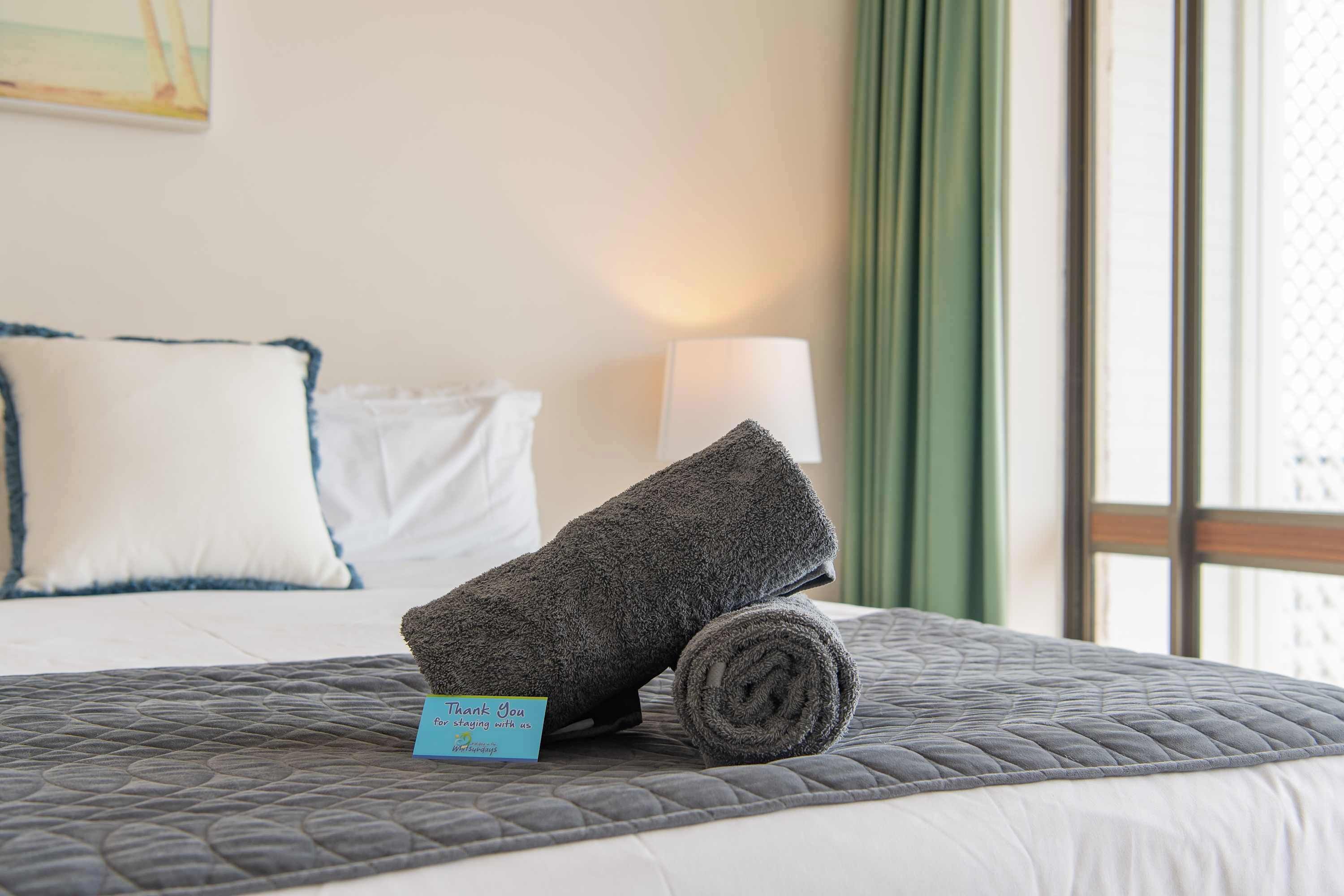 A neatly made bed with a rolled-up towel and a thank you card on a grey quilted bedspread.