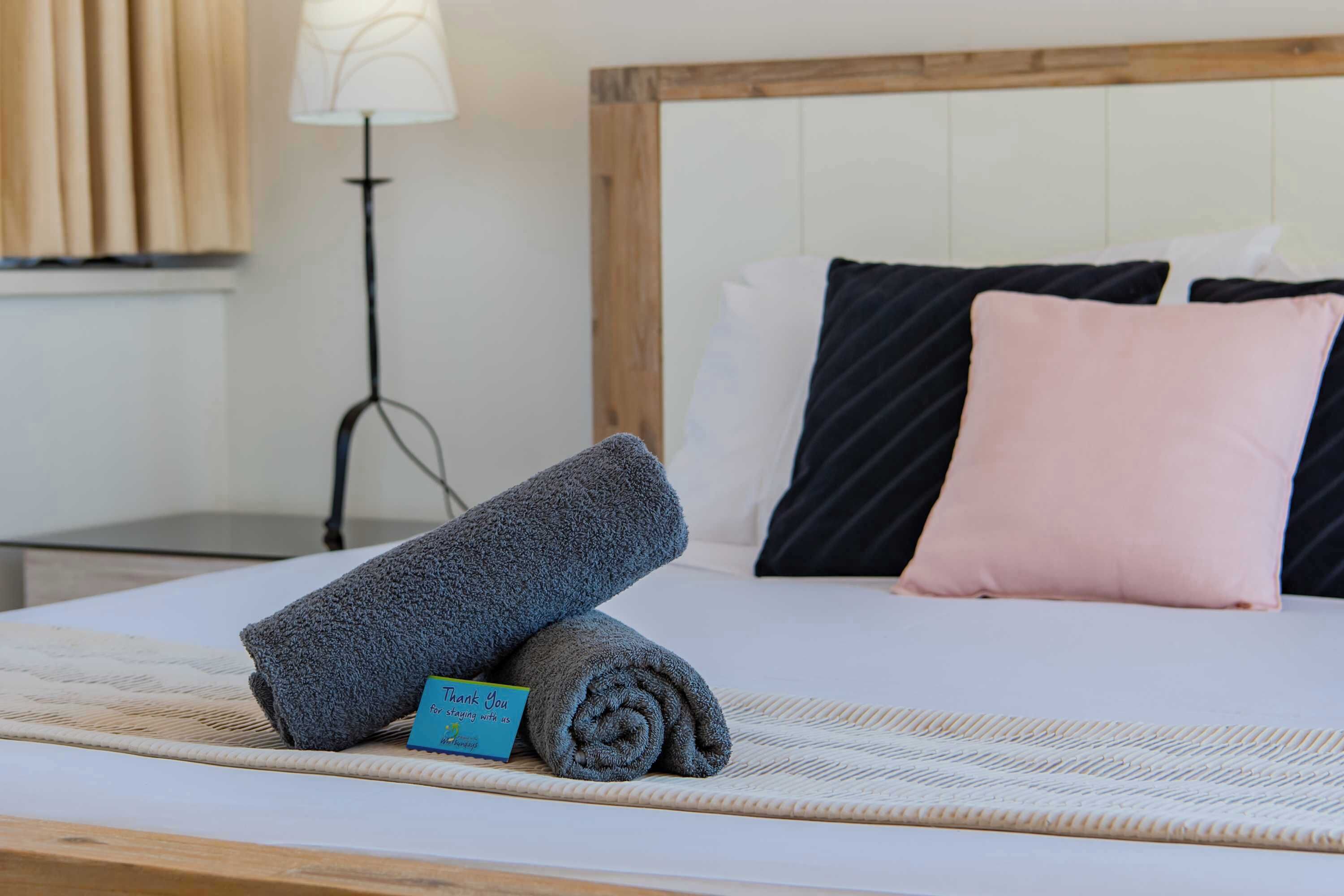 Un lit bien fait, avec des serviettes pliées en rouleau et une carte de remerciement, une lampe de chevet et une tête de lit en bois.