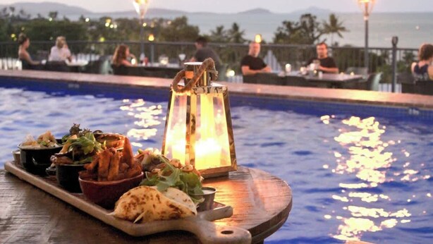 A dining area with a poolside view, featuring a wooden serving board with food and a candle. - OCEAN VIEWS 10A pool & Spa Wi-Fi (Airlie Beach)