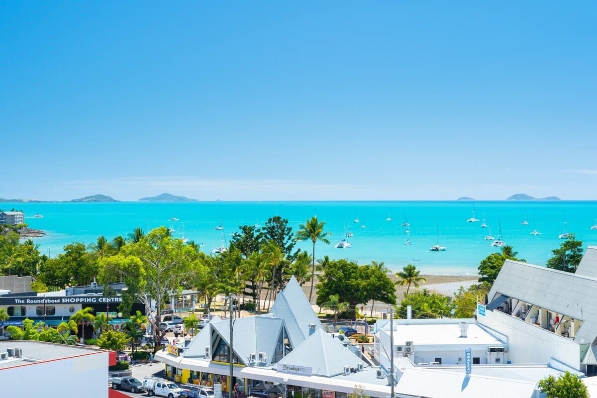 A coastal town with a prominent white building with a triangular roof, a visible sign for 'The Roundabout Shopping Centre', and a clear view of the turquoise sea with boats.